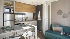 Eine kompakte Küche mit modernen Geräten, ein Essbereich mit weißem Tisch und Stühlen sowie ein Salontisch mit einem türkisfarbenen Sofa und einer Zeitschrift.