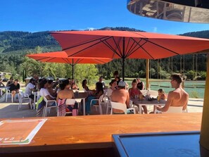 An outdoor dining area with tables and chairs, a large red umbrella, and a view of a lake and hills. - Chalet 4 étoiles - Piscine  - ccbgbad (Arques)