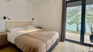 A modern bedroom with a large bed, a nightstand, a lamp, and a view of a body of water through a sliding glass door.