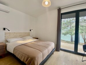 A modern bedroom with a large bed, a nightstand, a lamp, and a view of a body of water through a sliding glass door. - Villa 4 étoiles - Piscine  - ccbgbaf (Arques)