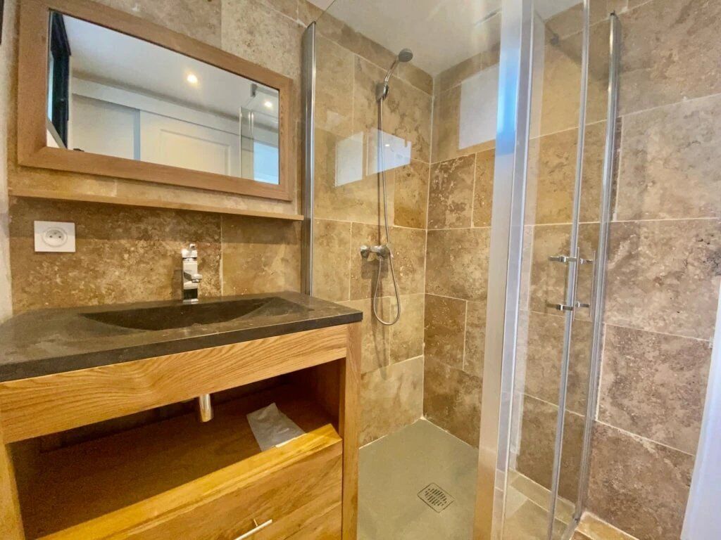 Een badkamer met een houten wastafelmeubel, een spiegel, een douche met glazen deuren en betegelde wanden.