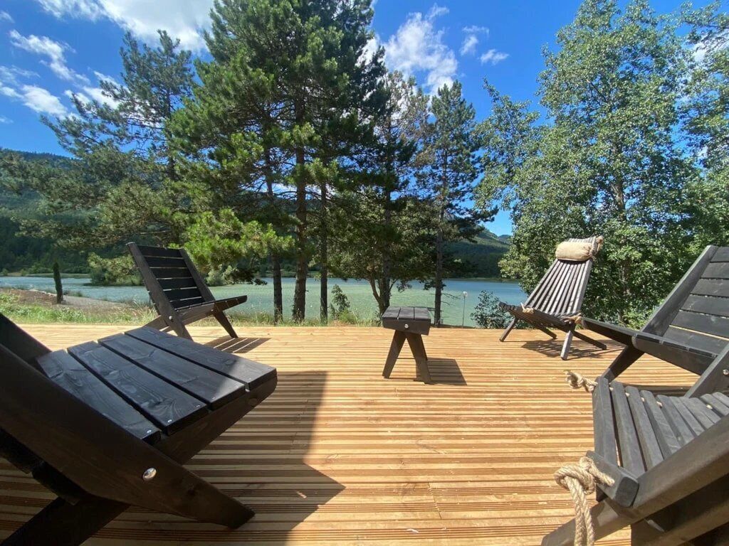 A wooden deck with lounge chairs and a small table, overlooking a lake and forested area.