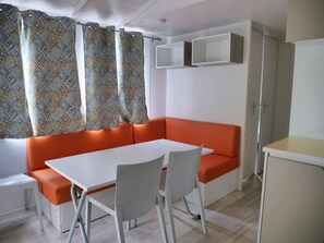 Ein gemütlicher Raum mit einem weißen Tisch, zwei Stühlen, einem orangefarbenen Sofa, einer kleinen Kommode und einem Fenster mit gemusterten Vorhängen.