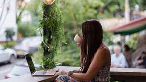 Una persona sentada a una mesa con un ordenador portátil y una taza, al aire libre, con follaje e iluminación colgante.