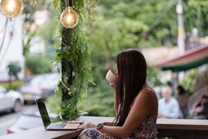 Una persona sentada a una mesa con un ordenador portátil y una taza, al aire libre, con follaje e iluminación colgante.