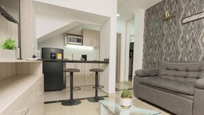 Un espacio de estar moderno con un sofá gris, una mesa de centro de vidrio y una cocina al fondo.