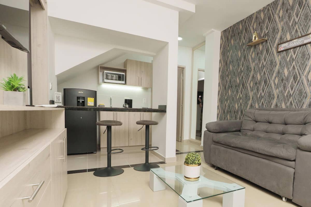 Ein moderner Wohnbereich mit grauem Sofa, einem Glastisch und einer Küche im Hintergrund.