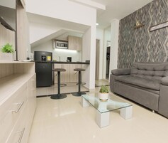 Un espacio de estar moderno con un sofá gris, una mesa de centro de vidrio y una cocina al fondo.