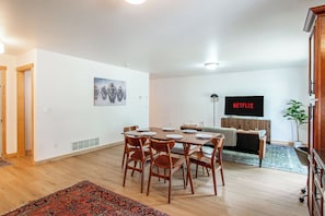 A modern living room with a dining area, a flat-screen TV, and a large window.