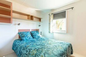 A bedroom with a bed, built-in shelves, a window with curtains, and a wall-mounted light fixture. - Camping 3 étoiles - ccbgcfd (Saint-Bonnet-près-Orcival)