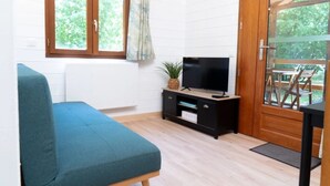 Ein Zimmer mit einem blauen Sofa, einem Fernseher auf einem Holzständer, einem Fenster mit Vorhängen und einer Tür zu einem Außenbereich.