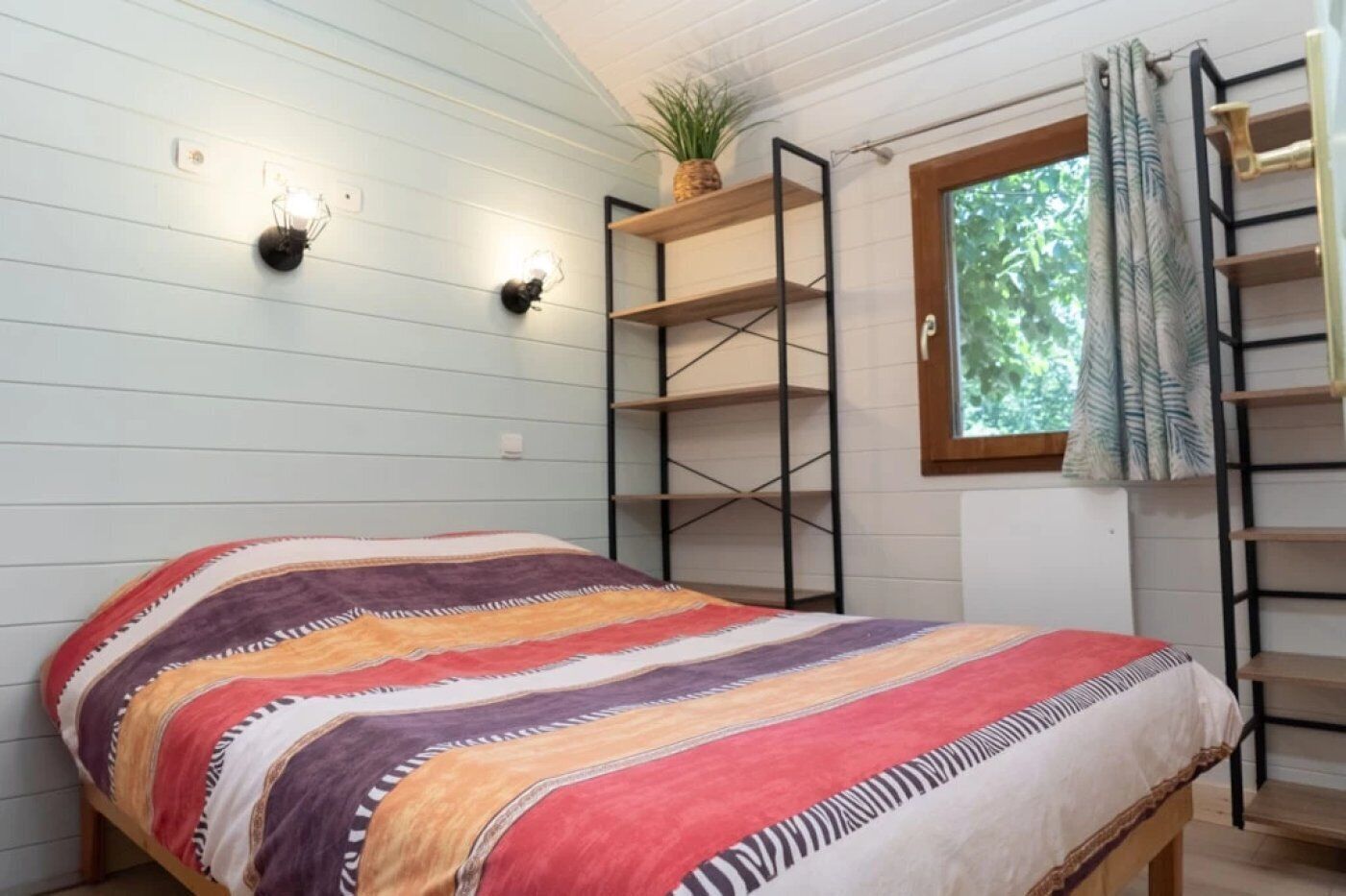 Een kamer met een bed, wandplanken, een raam met gordijnen en een plant.