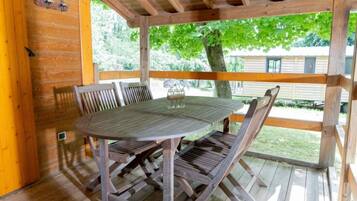 Een houten terras met een tafel, stoelen, een glas water en uitzicht op een huis en bomen.