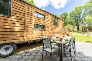 A wooden cabin with a patio, a table with chairs, and a window with a view of the surrounding greenery. - Camping 3 étoiles - ccbgcfh (Saint-Bonnet-près-Orcival)