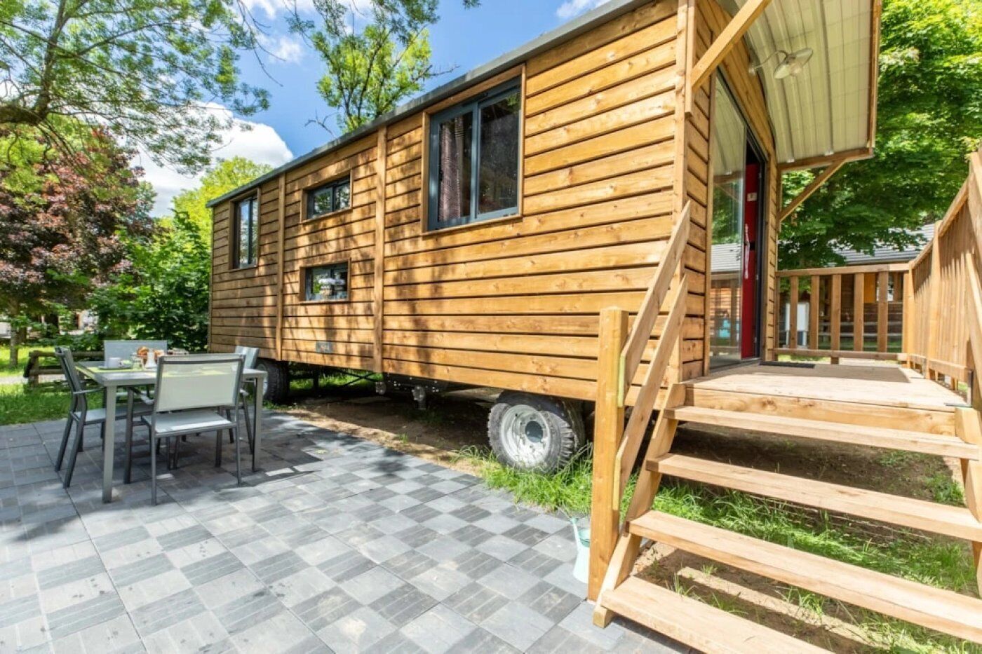 A wooden mobile home with a patio area, a dining table, and a staircase leading to the entrance.