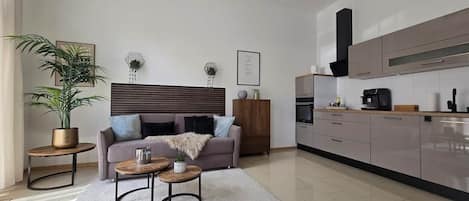 Una sala de estar moderna con un sofá, dos mesas auxiliares, una planta en maceta y una zona de cocina con armarios y electrodomésticos.
