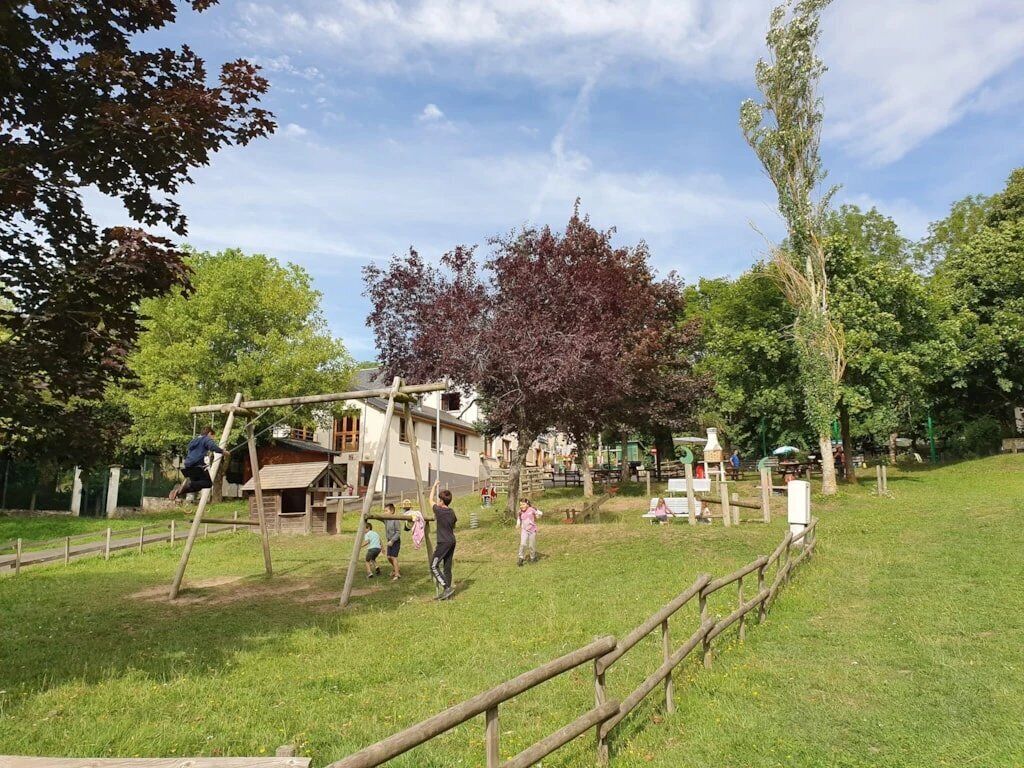 Une aire de jeux avec des structures en bois, une zone herbeuse et des arbres en arrière-plan.