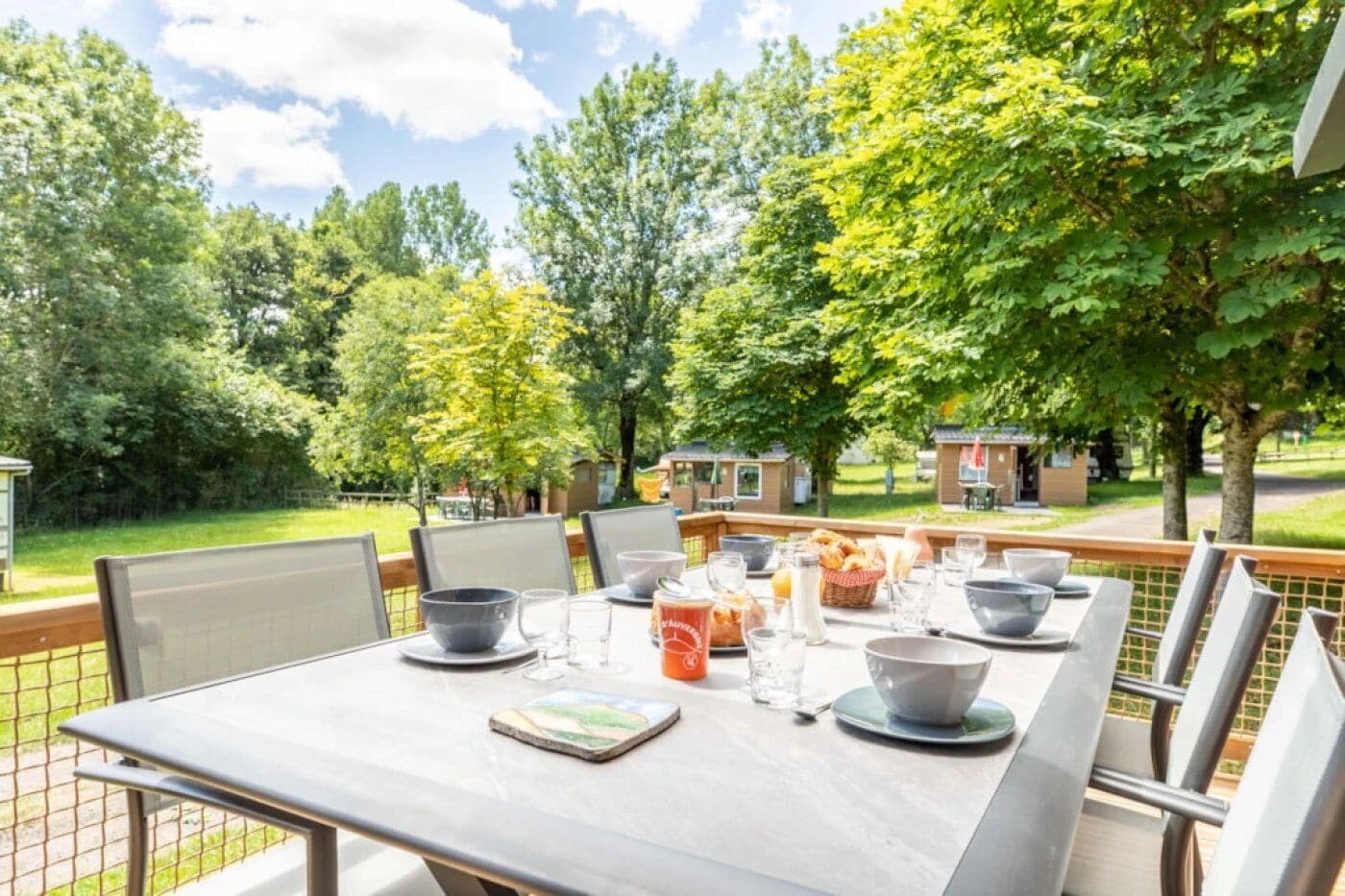 Un espace de restauration en plein air avec une table dressée pour quatre personnes, entourée de chaises, et offrant une vue sur un jardin et un bâtiment en arrière-plan.