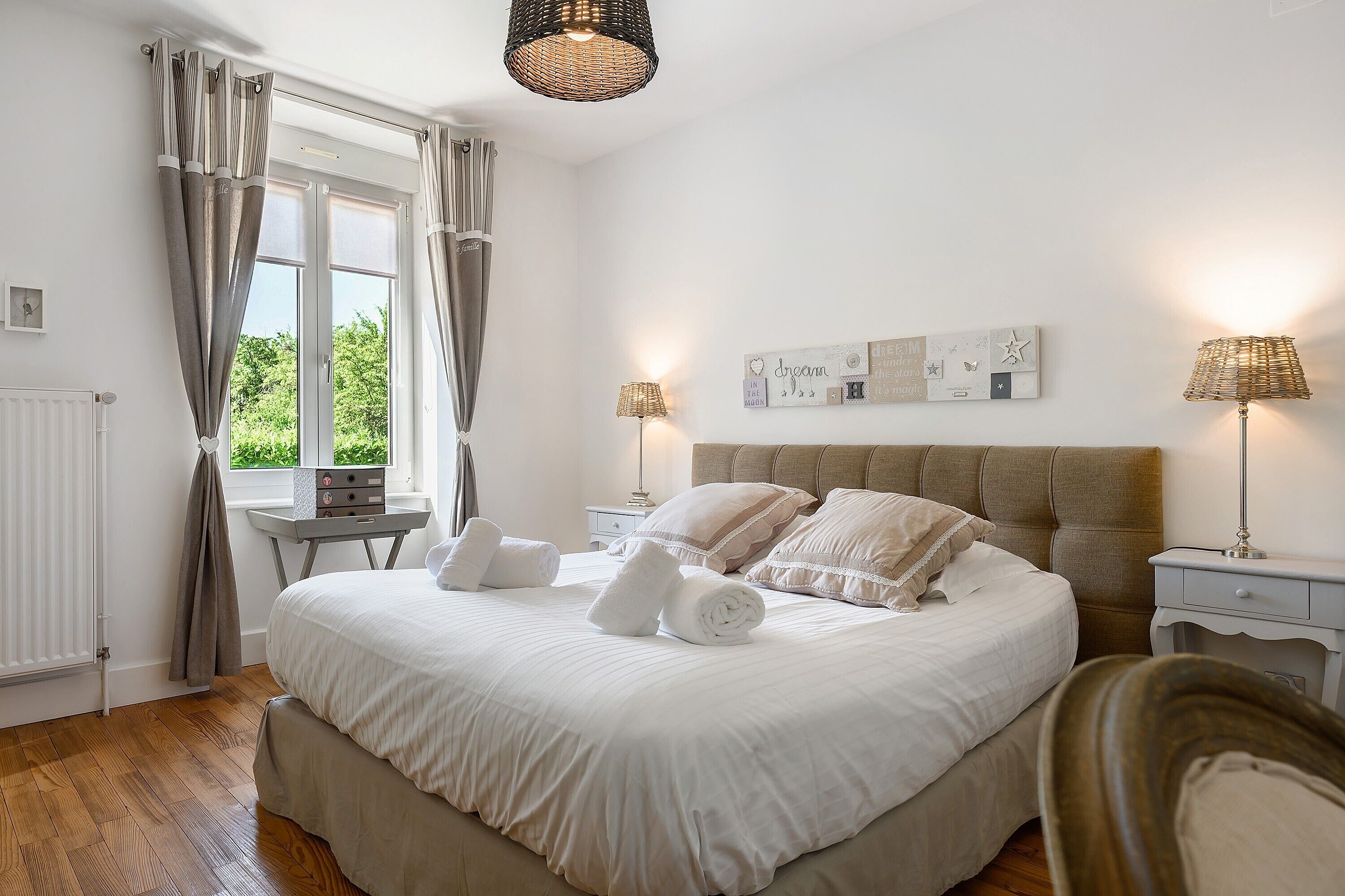Une chambre à coucher bien aménagée, avec un grand lit, deux tables de chevet, une commode, une fenêtre avec des rideaux et une œuvre d’art accrochée au mur.