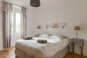 Ein Schlafzimmer mit einem Bett, zwei Nachttischen, einem Fenster mit Vorhängen, einem Wandbild und einer Pendelleuchte.