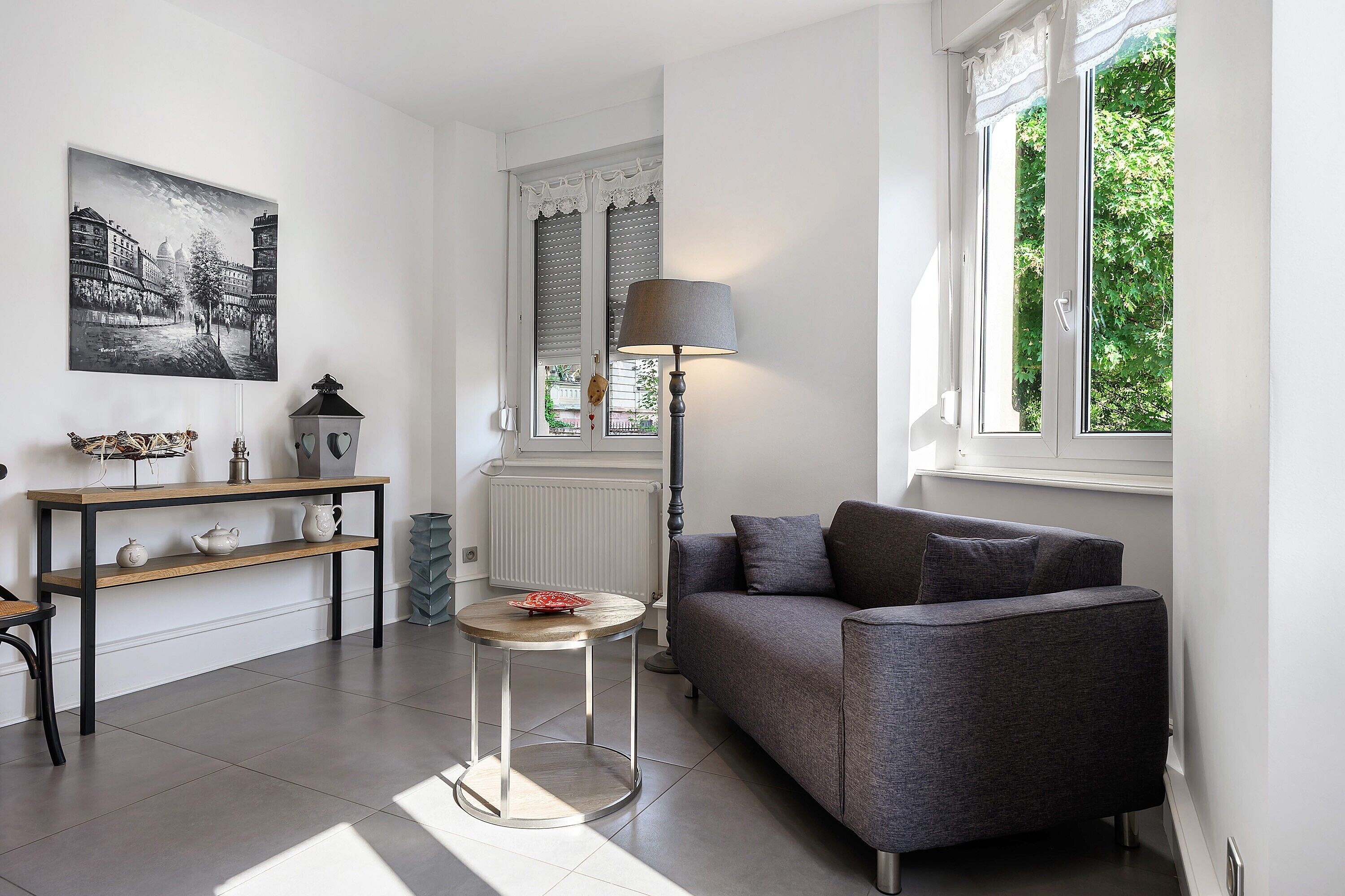 Ein modernes Wohnzimmer mit einem grauen Sofa, einem kleinen runden Tisch, einer Lampe und einer Heizung. An der Wand hängt ein Gemälde und das Fenster bietet Blick auf Grünflächen.