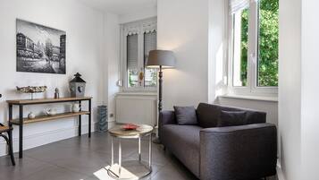 Ein modernes Wohnzimmer mit einem grauen Sofa, einem kleinen runden Tisch, einer Lampe und einer Heizung. An der Wand hängt ein Gemälde und das Fenster bietet Blick auf Grünflächen.