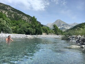 Ein klares Flussufer mit einer Person im Wasser, umgeben von felsigem Gelände und viel Grün.
