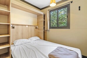 Un lit simple avec une literie blanche, une tête de lit en bois et des étagères intégrées sur le côté gauche. Une fenêtre donnant sur des arbres se trouve sur le côté droit.