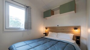 A bedroom with a bed, a window with curtains, and wall-mounted lighting. - Camping 3 étoiles - Piscine  - ccbgebh (Pianottoli-Caldarello)