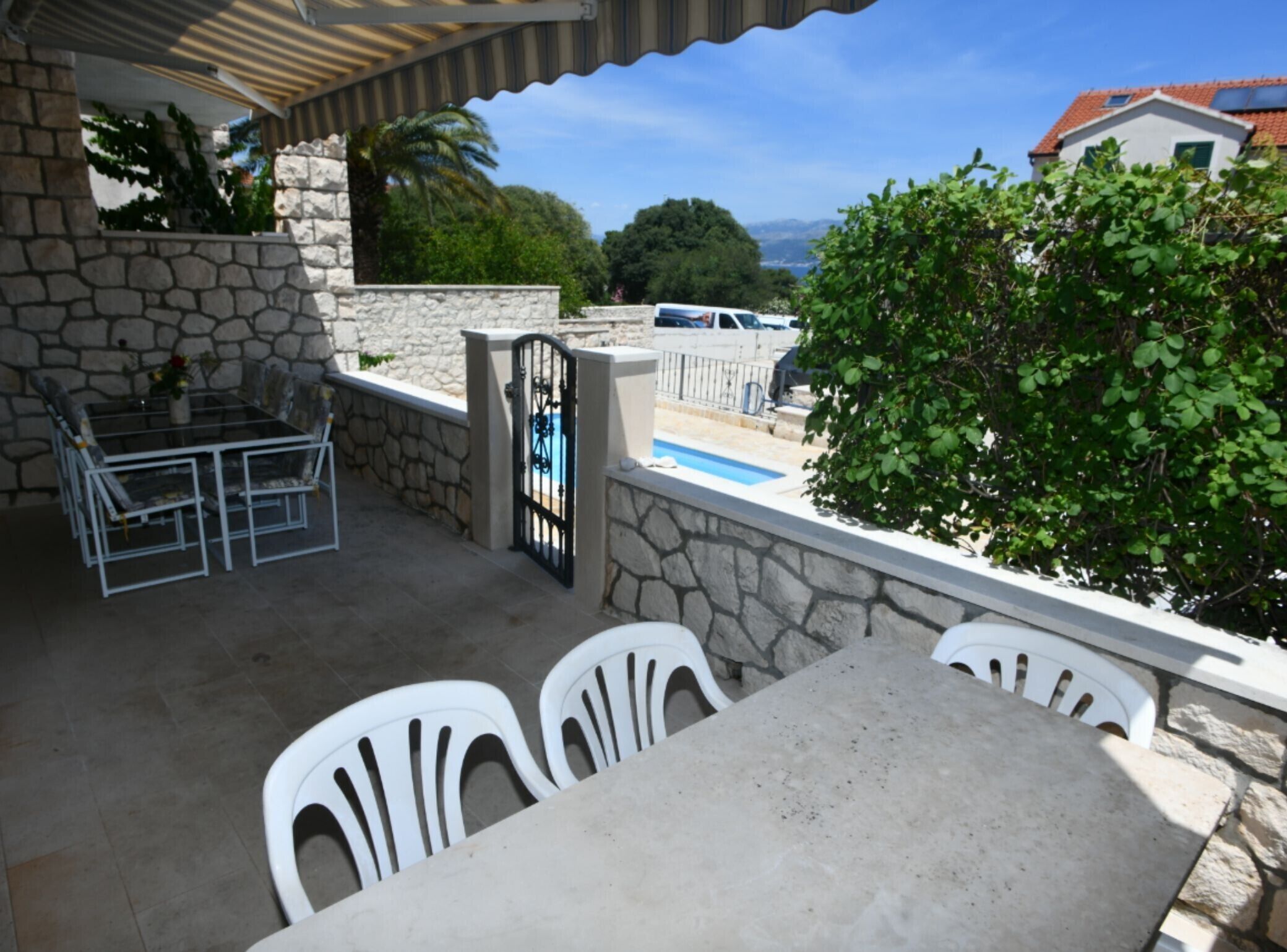 A patio with a table and chairs, a stone wall, a gate, and a view of the sea and buildings in the distance.
