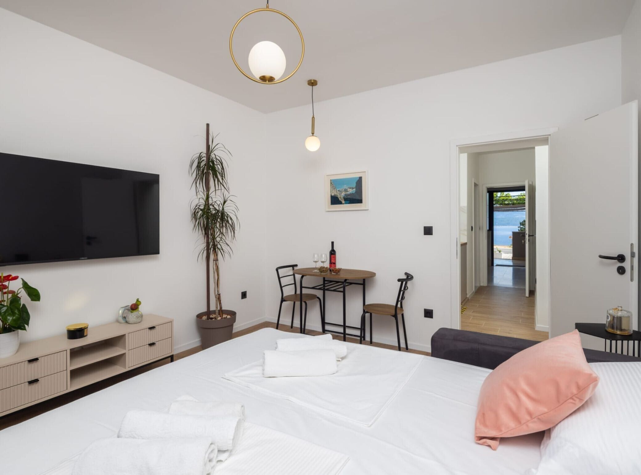 Une chambre moderne avec un lit, des tables de chevet, une télévision, un coin repas avec des chaises, une plante en pot et une vue sur l’extérieur.