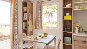 A dining area with a table, chairs, and a window with curtains. There are shelves with kitchen items and a view of trees outside. - Camping 3 étoiles - Piscine  - ccbgecb (Pianottoli-Caldarello)