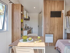 Ein gemütlicher Raum mit Esstisch aus Holz, einer kleinen Küchenzeile, einem Bett mit rosa Decke und einem an der Wand befestigten Fernseher.