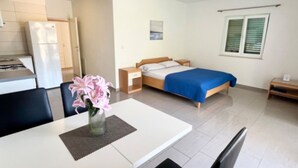 Una habitación de hotel moderna con una cama, mesita de noche y una pequeña mesa con sillas. Cuenta con un área de kitchenette con refrigerador y estufa.