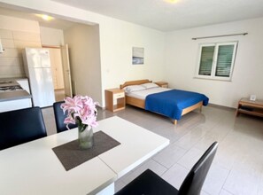 Ein modernes Hotelzimmer mit einem Bett, Nachttisch und einem kleinen Tisch mit Stühlen. Es gibt eine Küchenzeile mit Kühlschrank und Herd.