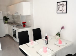 Eine moderne Küche mit weißen Schränken, einem Herd und einer Spüle. Ein Esstisch ist gedeckt mit rosa Servietten, Tellern und einer Weinflasche.