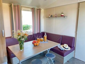 Une pièce avec une table en bois, des sièges rembourrés de couleur violette, un vase rempli de fleurs, un panier de fruits et une bouteille de jus.