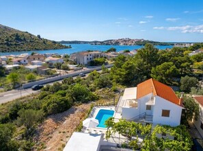 A coastal property with a swimming pool, surrounded by greenery and a view of the sea and a hill.