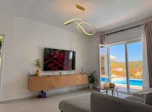 A modern living room with a flat-screen TV, a sofa, a dining table with candles, and a view of a swimming pool.