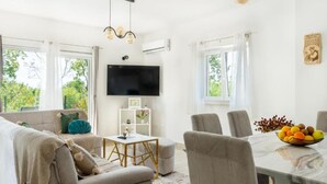 A modern living room with a sofa, a dining table with chairs, a TV, and a window with a view of greenery. - Villa with mountain view (Lovreć)