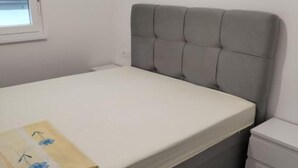 Ein Bett mit grau gepolstertem Kopfteil, einer weißen Matratze und einem kleinen Nachttisch mit Schubladen.