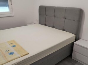 Ein Bett mit grau gepolstertem Kopfteil, einer weißen Matratze und einem kleinen Nachttisch mit Schubladen.