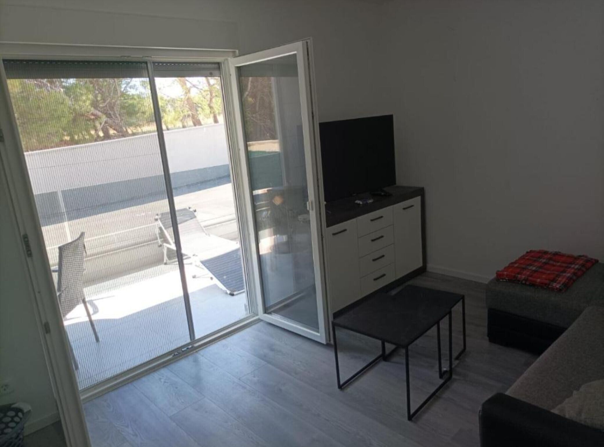 Et værelse med en skydedør af glas, der fører til en balkon, et fjernsyn på et skab, et lille bord og en sofa.
