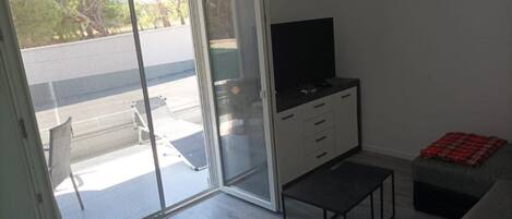 Et værelse med en skydedør af glas, der fører til en balkon, et fjernsyn på et skab, et lille bord og en sofa.