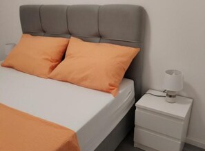 Ein Bett mit grauem Polsterkopfstück, zwei orangefarbenen Kissen und einem weißen Nachttisch mit einer Lampe.