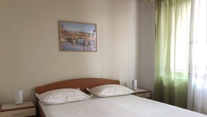 Un lit bien fait, avec du linge de lit blanc et deux oreillers, une tête de lit en bois, une table de chevet avec une lampe et un tableau représentant un pont accroché au mur.