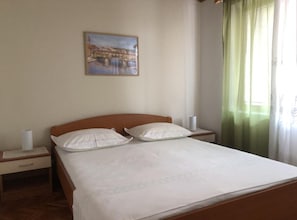 A neatly made bed with white linens and two pillows, a wooden headboard, a nightstand with a lamp, and a painting of a bridge on the wall. - Two Bedroom Apartment with Terrace (Drasnice)