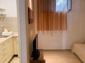 Un dormitorio con cama, televisor sobre un mueble de madera, ventana con cortinas y baño con lavamanos y toalla.