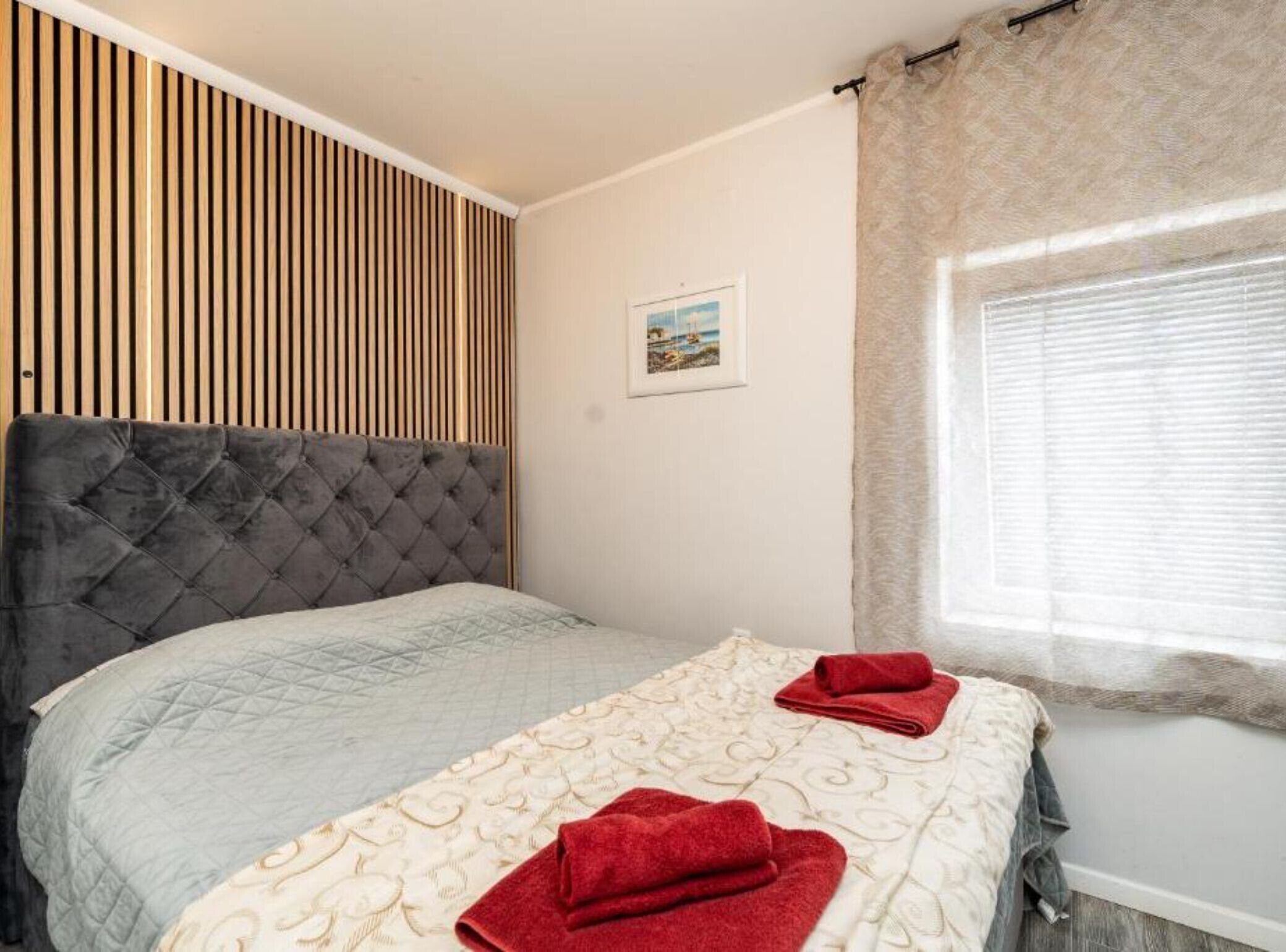 A bedroom with a tufted headboard, a bed with a quilted bedspread, two red towels, a window with curtains, and a wooden slat wall panel.