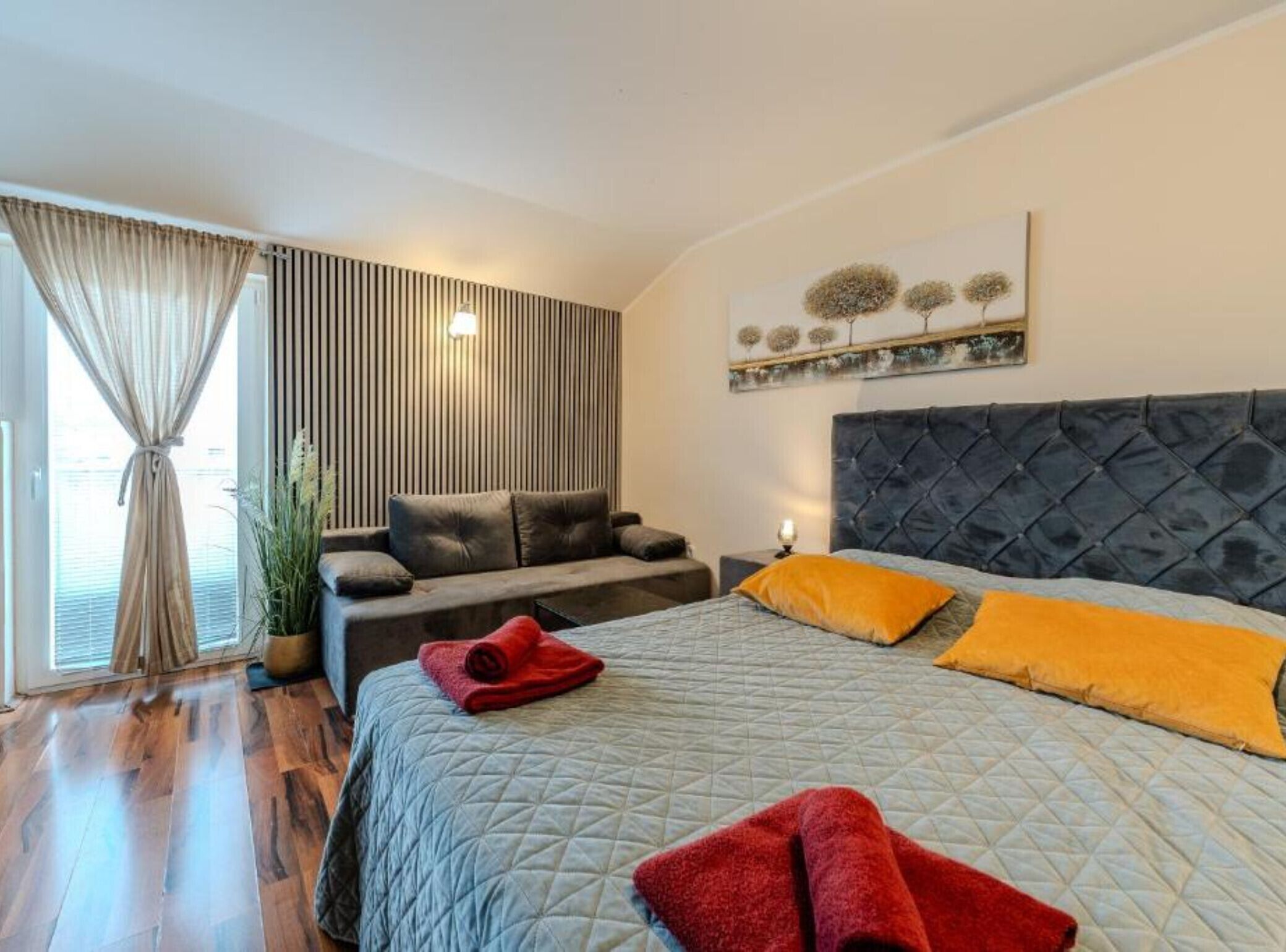 Une chambre à coucher avec un lit, un canapé, une fenêtre avec des rideaux et un mur orné d’une plante et d’un tableau.